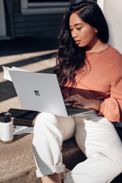Photo by Windows woman in orange long sleeve shirt and white pants sitting on floor using Surface Laptop